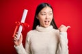 Young asian student woman holding graduate diploma over red isolated background pointing and showing with thumb up to the side Royalty Free Stock Photo