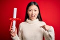 Young asian student woman holding graduate diploma over red isolated background with surprise face pointing finger to himself Royalty Free Stock Photo