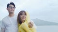 Young Asian sweet couple feeling happy using romantic time playing rain while wearing raincoat standing near lake. Lifestyle