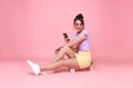 Young asian teenage girl sitting on skateboard and holding smartphone on pink background