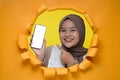 Young Asian teenager girl poses through torn yellow paper hole, showing mobile phone with blank white screen display for a copy Royalty Free Stock Photo