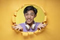 Young Asian teenager man confused with spread hands poses through torn yellow paper hole, wearing purple t-shirt and headphone Royalty Free Stock Photo