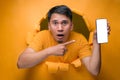 Young Asian teenager man with shock face poses through torn yellow paper hole, showing mobile phone with blank white screen Royalty Free Stock Photo