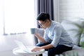 Young Asian wear glasses, use a laptop and hold a hot coffee cup and sit on the sofa near the large window Royalty Free Stock Photo