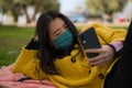 Young Asian woman in city park during covid19 - happy and pretty Japanese girl in face mask using internet with mobile phone Royalty Free Stock Photo