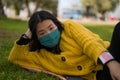 Young Asian woman in city park during covid19 - outdoors lifestyle portrait of happy and pretty Japanese girl in face mask lying Royalty Free Stock Photo