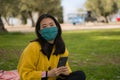 Young Asian woman in city park during covid19 - outdoors lifestyle portrait of happy and pretty Japanese girl in face mask using