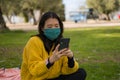 Young Asian woman in city park during covid19 - outdoors lifestyle portrait of happy and pretty Korean girl in face mask using Royalty Free Stock Photo