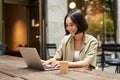 Young asian woman, digital nomad working remotely from a cafe, drinking coffee and using laptop, smiling Royalty Free Stock Photo