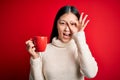 Young asian woman drinking a cup of hot coffee over isolated red background with happy face smiling doing ok sign with hand on eye Royalty Free Stock Photo