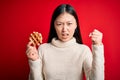 Young asian woman eating sweet and tasty belgian waffle over red isolated background annoyed and frustrated shouting with anger,