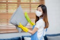 Young asian woman in face mask and gloves cleaning dust with duster on sofa and cushions at home.