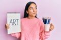 Young asian woman holding art notebook and colored pencils smiling looking to the side and staring away thinking Royalty Free Stock Photo