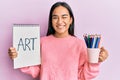 Young asian woman holding art notebook and colored pencils winking looking at the camera with sexy expression, cheerful and happy Royalty Free Stock Photo