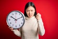 Young asian woman holding countdown big clock over red isolated background annoyed and frustrated shouting with anger, crazy and