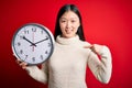 Young asian woman holding countdown big clock over red isolated background with surprise face pointing finger to himself Royalty Free Stock Photo