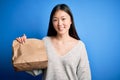 Young asian woman holding delivery paper bag for takeaway food over blue isolated background with a happy face standing and Royalty Free Stock Photo