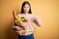 Young asian woman holding paper bag of fresh healthy groceries over yellow  background Pointing down looking sad and Royalty Free Stock Photo