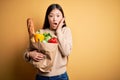 Young asian woman holding paper bag of fresh healthy groceries over yellow isolated background afraid and shocked, surprise and Royalty Free Stock Photo