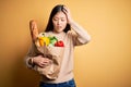 Young asian woman holding paper bag of fresh healthy groceries over yellow isolated background Looking at the watch time worried,