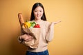 Young asian woman holding paper bag of fresh healthy groceries over yellow isolated background smiling cheerful presenting and Royalty Free Stock Photo