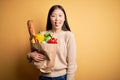 Young asian woman holding paper bag of fresh healthy groceries over yellow isolated background sticking tongue out happy with Royalty Free Stock Photo