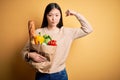 Young asian woman holding paper bag of fresh healthy groceries over yellow isolated background Strong person showing arm muscle, Royalty Free Stock Photo