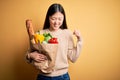 Young asian woman holding paper bag of fresh healthy groceries over yellow isolated background very happy and excited doing winner Royalty Free Stock Photo