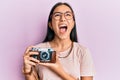 Young asian woman holding vintage camera angry and mad screaming frustrated and furious, shouting with anger looking up Royalty Free Stock Photo