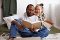 Young asian woman hugging african american boyfriend reading book near laptop on bed at home Royalty Free Stock Photo