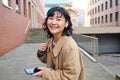 Young asian woman listens music in wireless earphones, uses smartphone on street, walks around city on sunny day Royalty Free Stock Photo