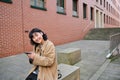 Young asian woman listens music in wireless earphones, uses smartphone on street, walks around city on sunny day Royalty Free Stock Photo