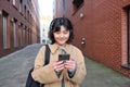 Young asian woman listens music in wireless earphones, uses smartphone on street, walks around city on sunny day Royalty Free Stock Photo