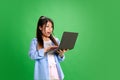 Young Asian woman looking to digital laptop with shocked facial expression against vibrant green studio background. Royalty Free Stock Photo