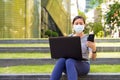 Young Asian woman with mask using phone and laptop while sitting on the stairs outdoors Royalty Free Stock Photo