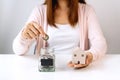 Young Asian woman`s hand putting coin in glass jar with model home on white table background. Saving, collect money for future, Royalty Free Stock Photo