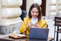 Young Asian woman sitting in coffee shop at wooden table, drinking coffee and using smartphone.On table is laptop. Girl browsing Royalty Free Stock Photo