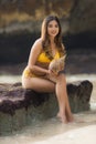 Young Asian woman sitting on the rock, holding coconuts. Summer beach concept. Tanned skin. Beautiful woman wearing yellow Royalty Free Stock Photo