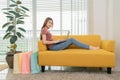 Young asian woman sitting on yellow sofa using laptop  shopping online near window and shopping bag Royalty Free Stock Photo