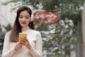 Young Asian woman smiling looking at mobile phone wearing headphones and long sleeves sweater while walking down the street Royalty Free Stock Photo