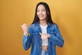 Young asian woman standing over yellow background pointing to the back behind with hand and thumbs up, smiling confident Royalty Free Stock Photo