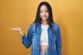 Young asian woman standing over yellow background smiling cheerful presenting and pointing with palm of hand looking at the camera Royalty Free Stock Photo