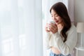 Young asian woman standing beside window and holding mug in bedroom at home Royalty Free Stock Photo