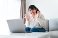 Young Asian woman using laptop computer for online video conference call waving hand making hello gesture on the couch in living Royalty Free Stock Photo