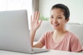 Young Asian woman using laptop computer for online video conference call waving hand making hello gesture in the living room Royalty Free Stock Photo
