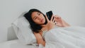 Young Asian woman using smartphone checking social media feeling happy smiling while lying on bed. Royalty Free Stock Photo