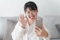 Young Asian woman using smartphone for online video conference call waving hand making hello gesture on the couch in living room Royalty Free Stock Photo