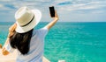 Young Asian woman wear straw hat in casual style use smartphone taking selfie at wooden pier. Summer vacation at tropical paradise