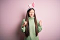 Young asian woman wearing cute easter bunny ears over pink background excited for success with arms raised and eyes closed