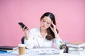 Young Asian woman wearing eyeglasses and a white shirt sits at a white desk with a laptop. She uses her smartphone to type sms Royalty Free Stock Photo